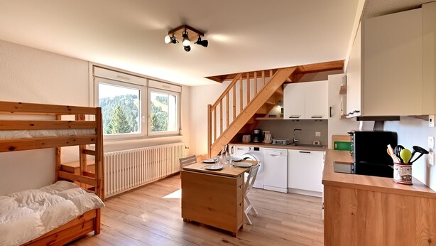 A cozy room with a bunk bed, wooden desk, and a kitchen area with white cabinets and a wooden staircase. - Le Nid des Vosges - Appt vue montagne (Gérardmer)