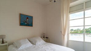 A neatly made bed with white linens, a painting of a sailboat on the wall, a window with a view of a river, and a nightstand with a lamp.