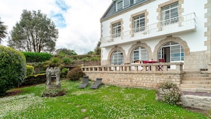Ein zweistöckiges Haus mit einer Steinaußenfassade, einem Balkon mit roten Stühlen und einem gepflegten Garten mit weißen Blumen und grünem Rasen.