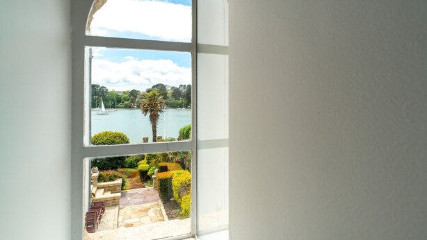 A view through a window with a stone archway, a body of water, and a landscaped garden with a palm tree and shrubs. - La Timonerie - Studio climatisé à Bénodet (Benodet)