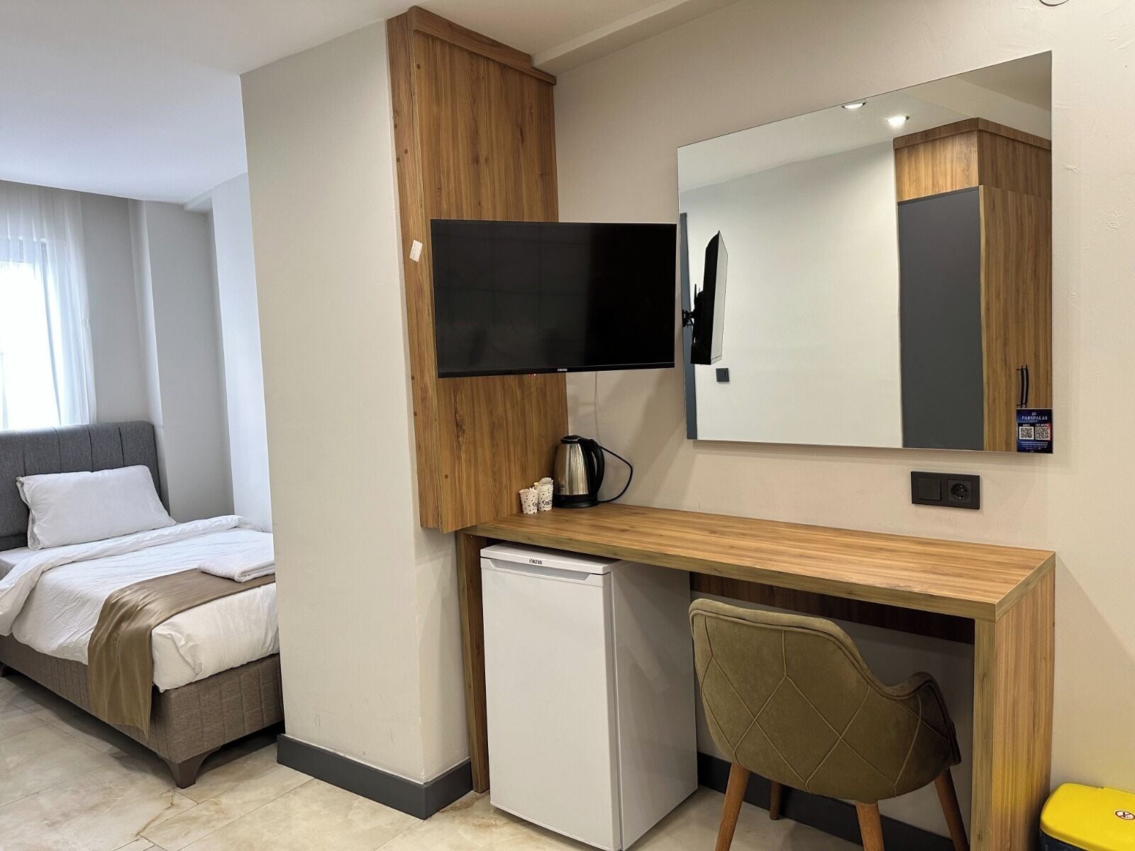 A hotel room with a bed, a desk with a computer, a small refrigerator, a chair, and a TV mounted on the wall.