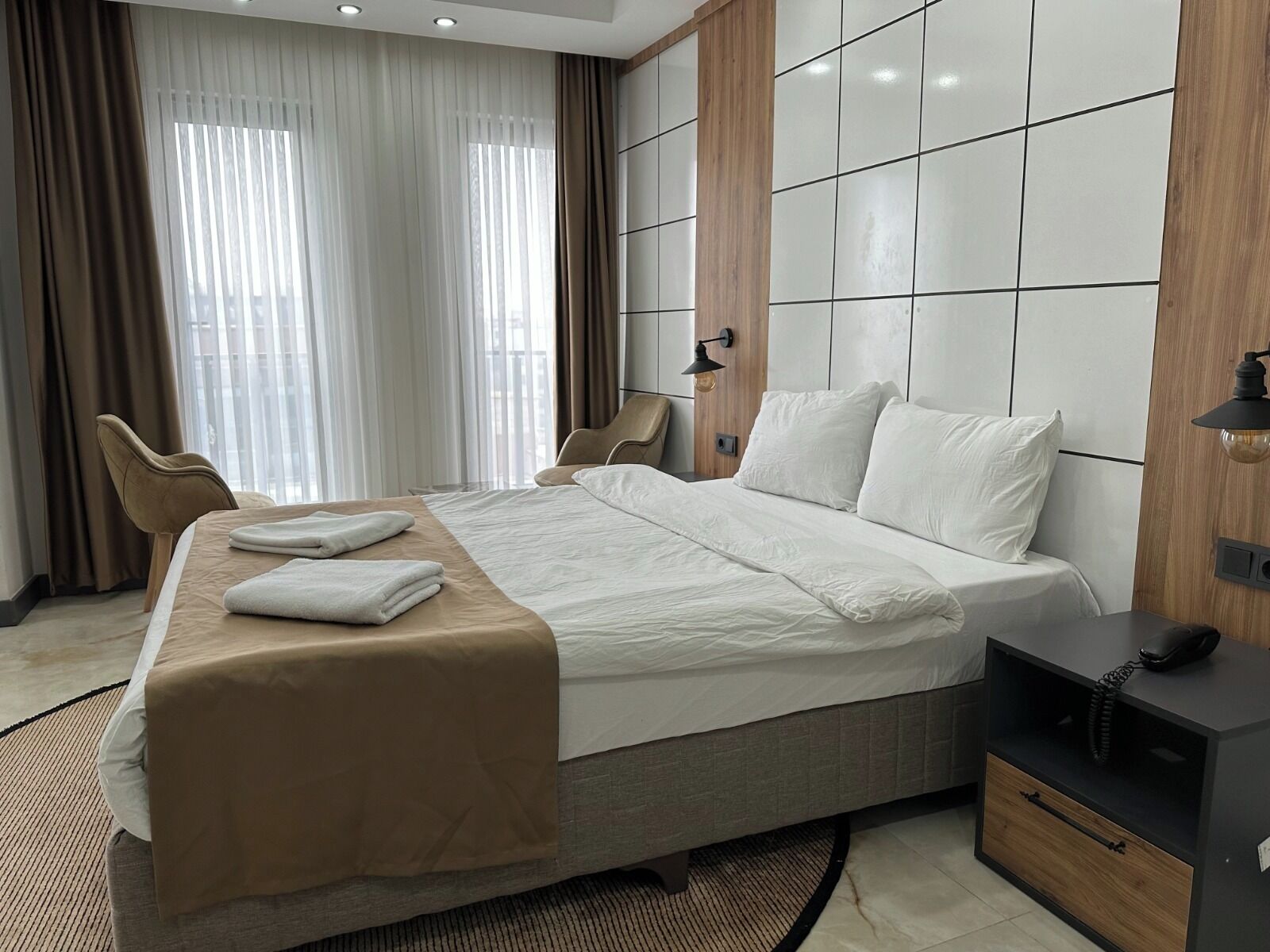 A modern hotel room with a large bed, two chairs, a bedside table with a lamp, and a view of the city through large windows.