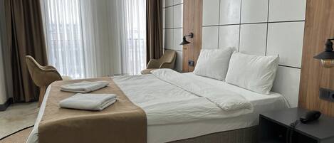 A modern hotel room with a large bed, two chairs, a bedside table with a lamp, and a view of the city through large windows.