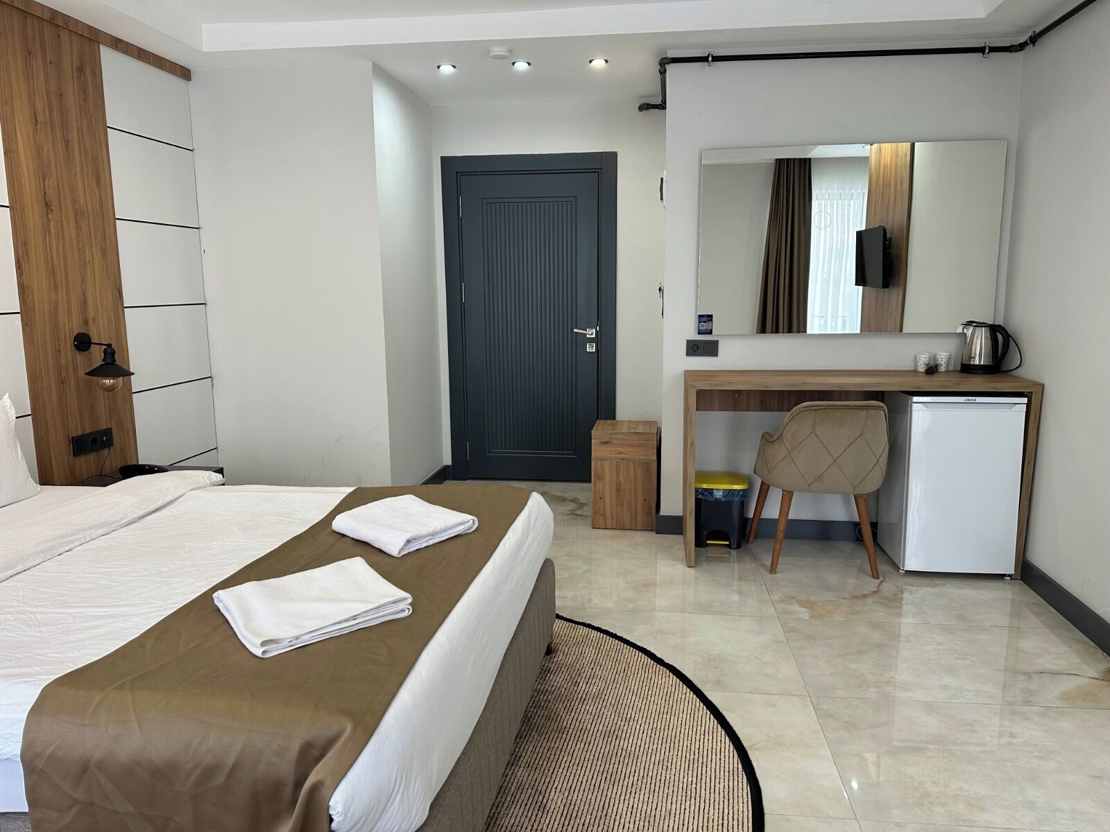 A hotel room with a bed, a desk, a chair, a small refrigerator, and a mirror.