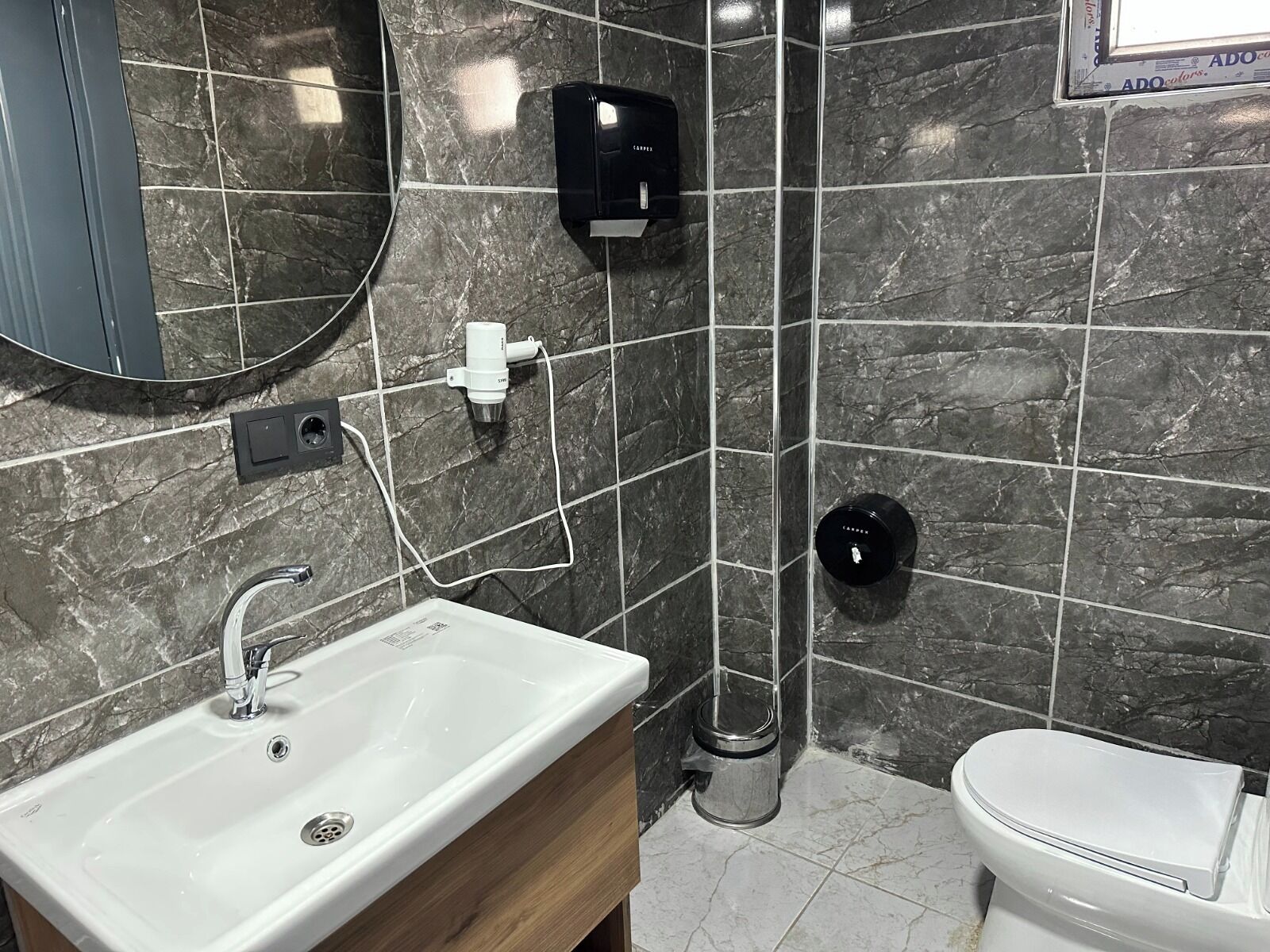 Une salle de bain avec un lavabo blanc, des toilettes, un miroir et un sèche-mains fixé au mur.