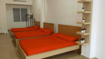 A room with three beds, each with orange bedding, a wooden headboard, and a white wall. There is a white air conditioning unit on the wall, a window with blinds, and a small shelf with potted plants.