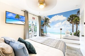 A bedroom with a large bed, a TV on the wall, and a view of the beach through an open door. - Coastal Oasis/4 BR/4 Mi to the Beach/Mini Golf/Game Room/Htd Pool/BBQ/Baby items (Hollywood)