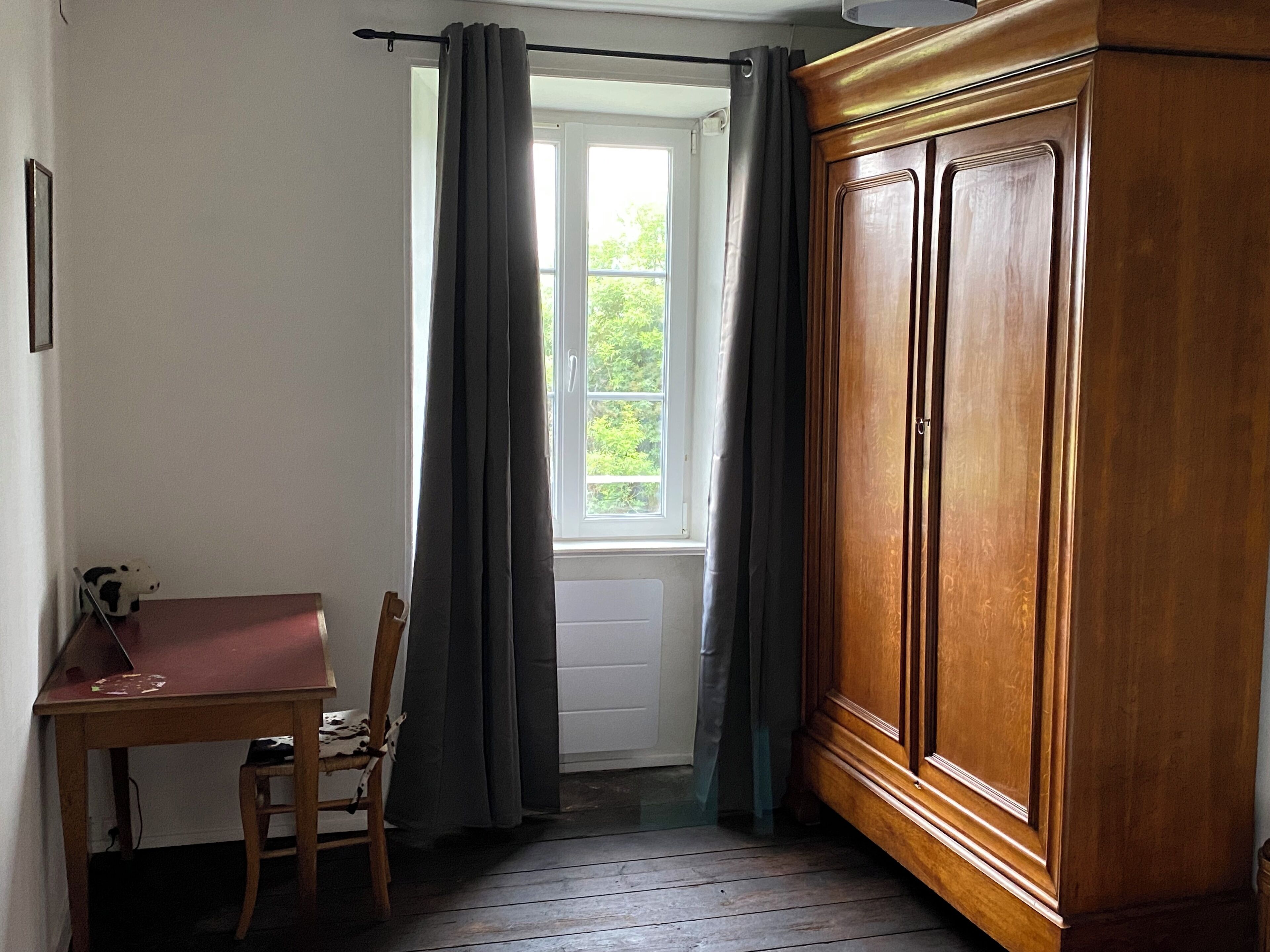 A room with a wooden wardrobe, a table with a chair, and a window with curtains.