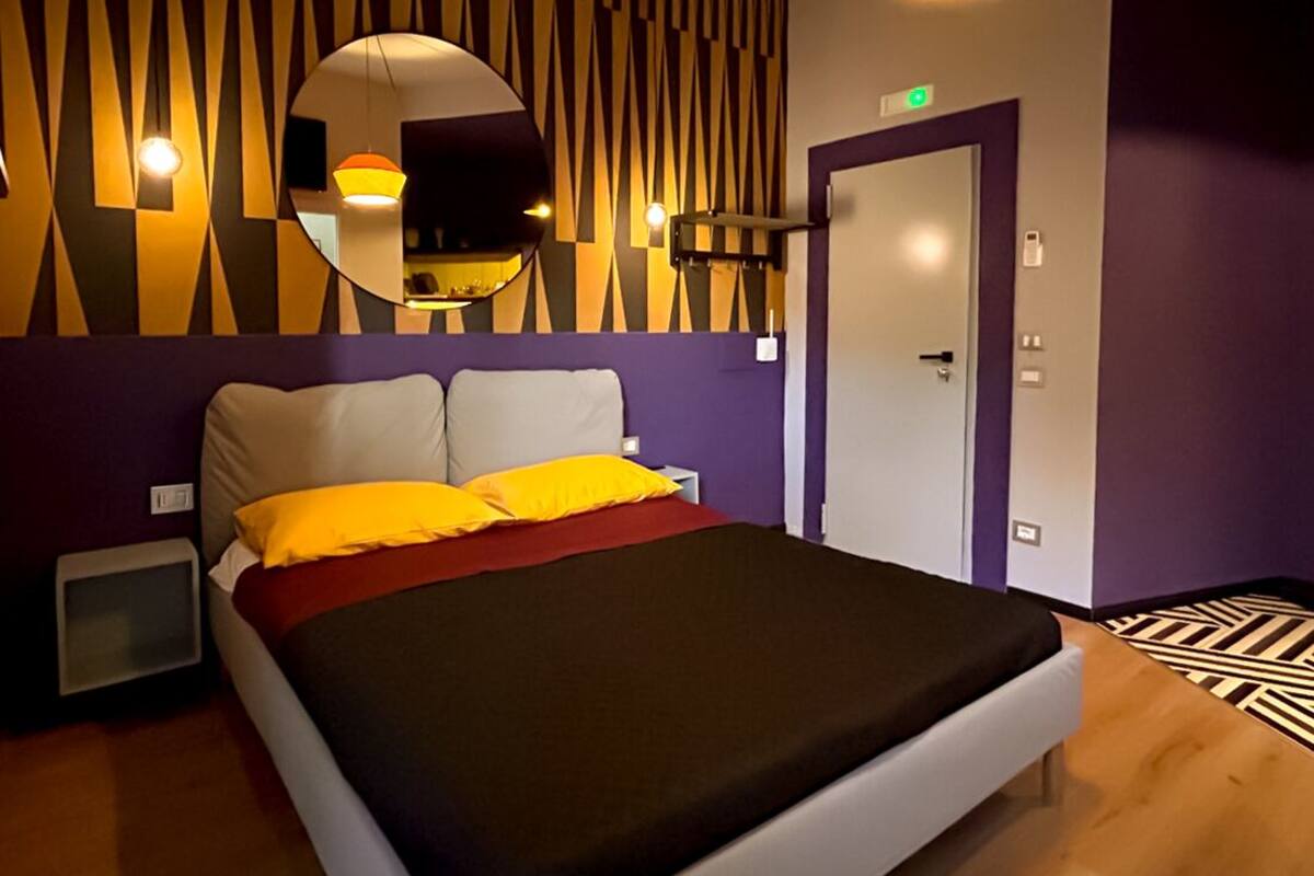 A modern hotel room with a bed, a mirror, a wall-mounted light fixture, and a door.