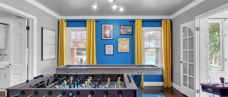 A room with a foosball table, a blue accent wall, yellow curtains, and a ceiling light fixture.