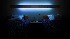 Une chambre d’hôtel à la lumière tamisée, avec deux lits, une table de chevet et un luminaire fixé au mur.