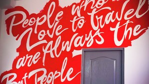 Une pièce dont un mur est décoré d’une fresque rouge sur laquelle on peut lire : « Les personnes qui aiment voyager sont toujours les meilleures », et une porte bleue est fermée.