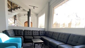 Un salon moderne avec un canapé d’angle, deux fauteuils et une table basse. On aperçoit un escalier au fond, qui mène à un niveau supérieur.