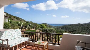 Un balcón con sofá, mesa y sillas, con vistas a un paisaje pintoresco con colinas y el mar.