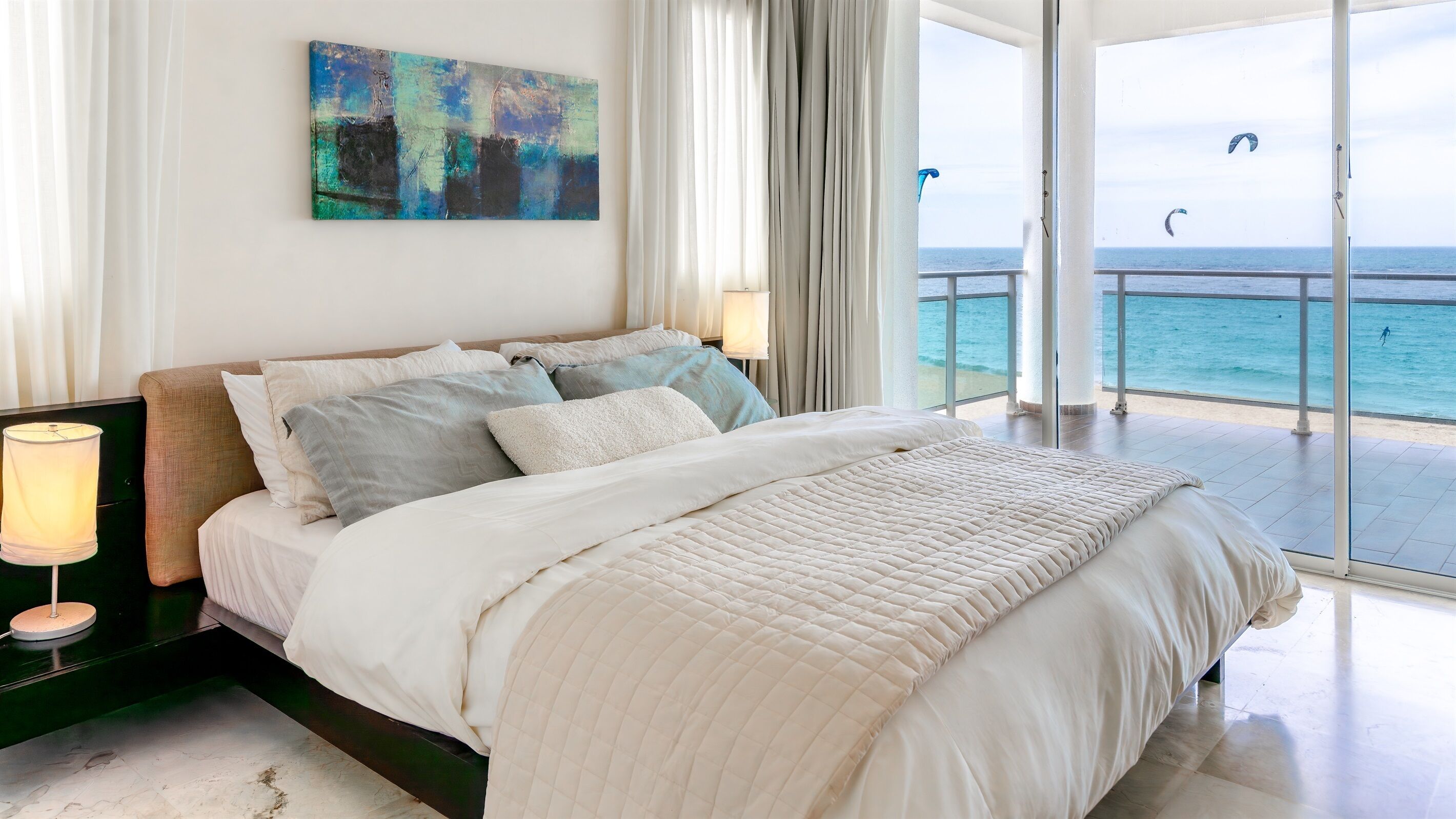 A bedroom with a large bed, a painting on the wall, and a view of the sea through the balcony doors.