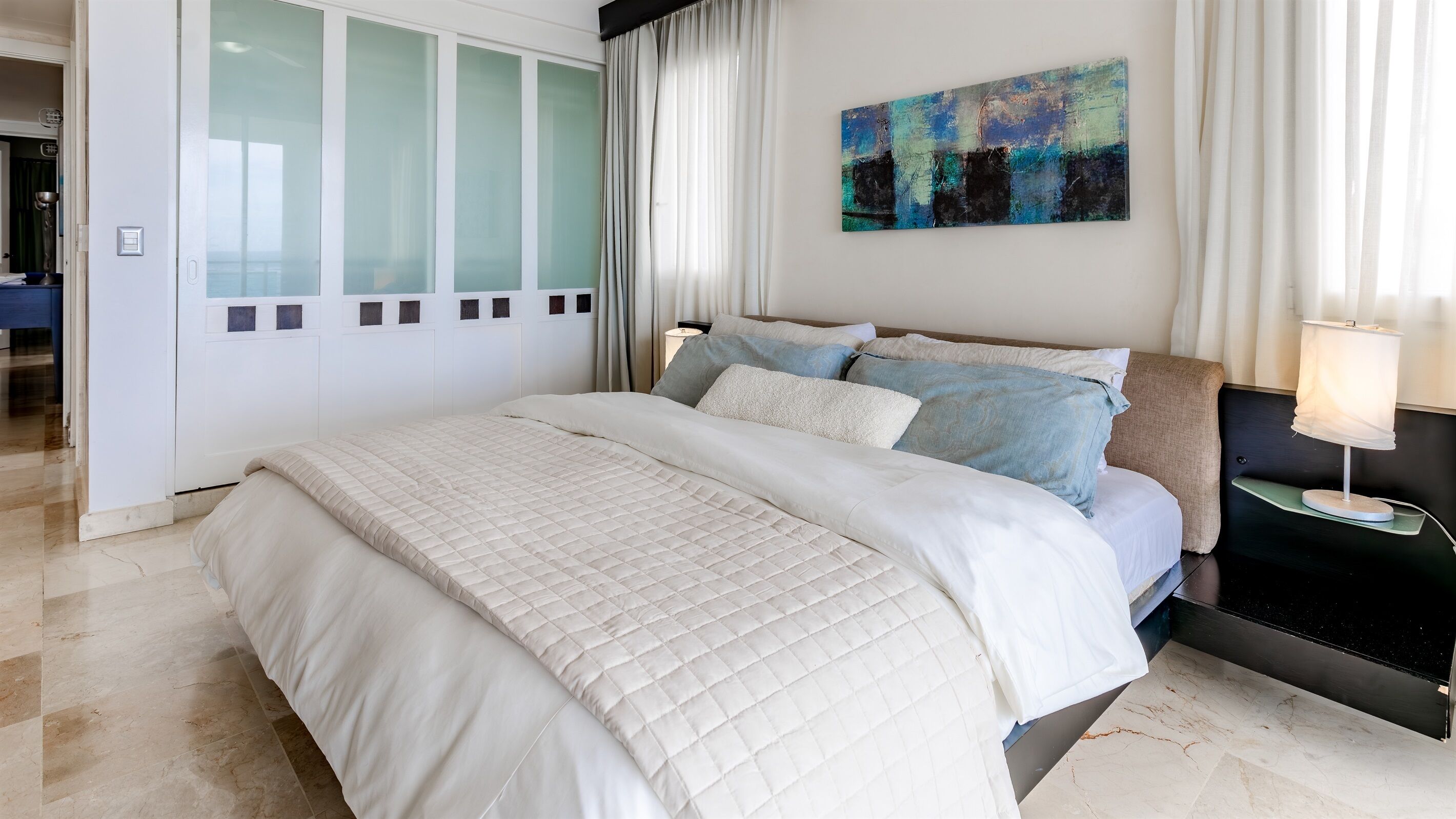 A modern bedroom with a large bed, a nightstand, a painting, and a view of the outside.