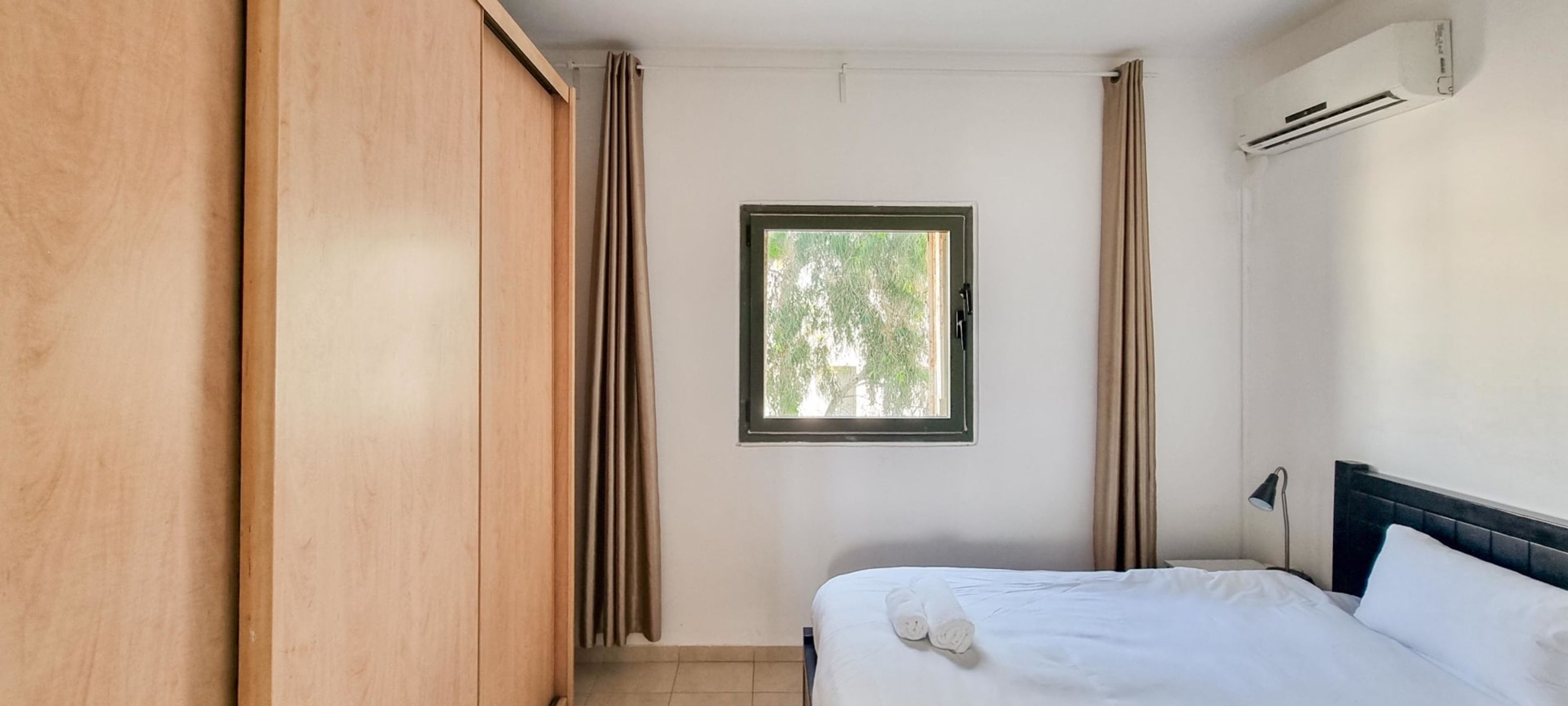 A bedroom with a bed, a window with curtains, an air conditioning unit, and a wardrobe.