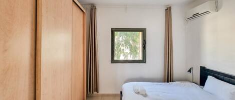 A bedroom with a bed, a window with curtains, an air conditioning unit, and a wardrobe.