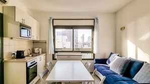 A compact living space with a kitchenette, a dining table, a sofa, and a large window offering a city view. - Ben Yehouda Obsidian (Tel Aviv)