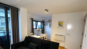 A modern living room with a black sofa, a dining table with chairs, and a large window with curtains.