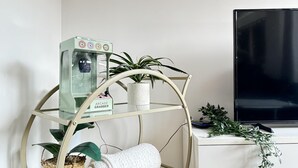 A room with a three-tiered metal shelf unit holding a water dispenser, a plant, and a rolled towel. A flat-screen TV is on a white cabinet to the right.