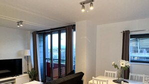 A modern living room with a black sofa, a dining table with white chairs, a TV, and a view of the city through large windows.