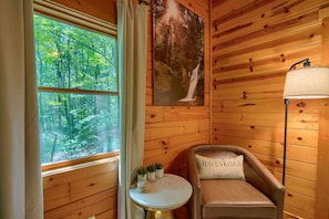 Interior acogedor de una cabaña con paredes de madera, una ventana con vista al bosque, una pequeña mesa con plantas, un sillón de cuero con cojín y una lámpara fijada a la pared.