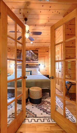 A wooden cabin interior with a bed, two ottomans, a patterned rug, and a ceiling fan. - 5 Min Old Mans Cave & Falls | Hot Tub | Game Table (Logan)