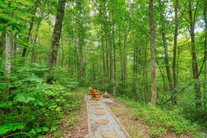 A forested area with a stone pathway leading to a wooden chair and a fire pit. - 5 Min Old Mans Cave & Falls | Hot Tub | Game Table (Logan)