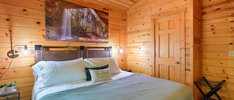 A wooden cabin bedroom with a bed, pillows, a nightstand, a wall-mounted picture, and a door.