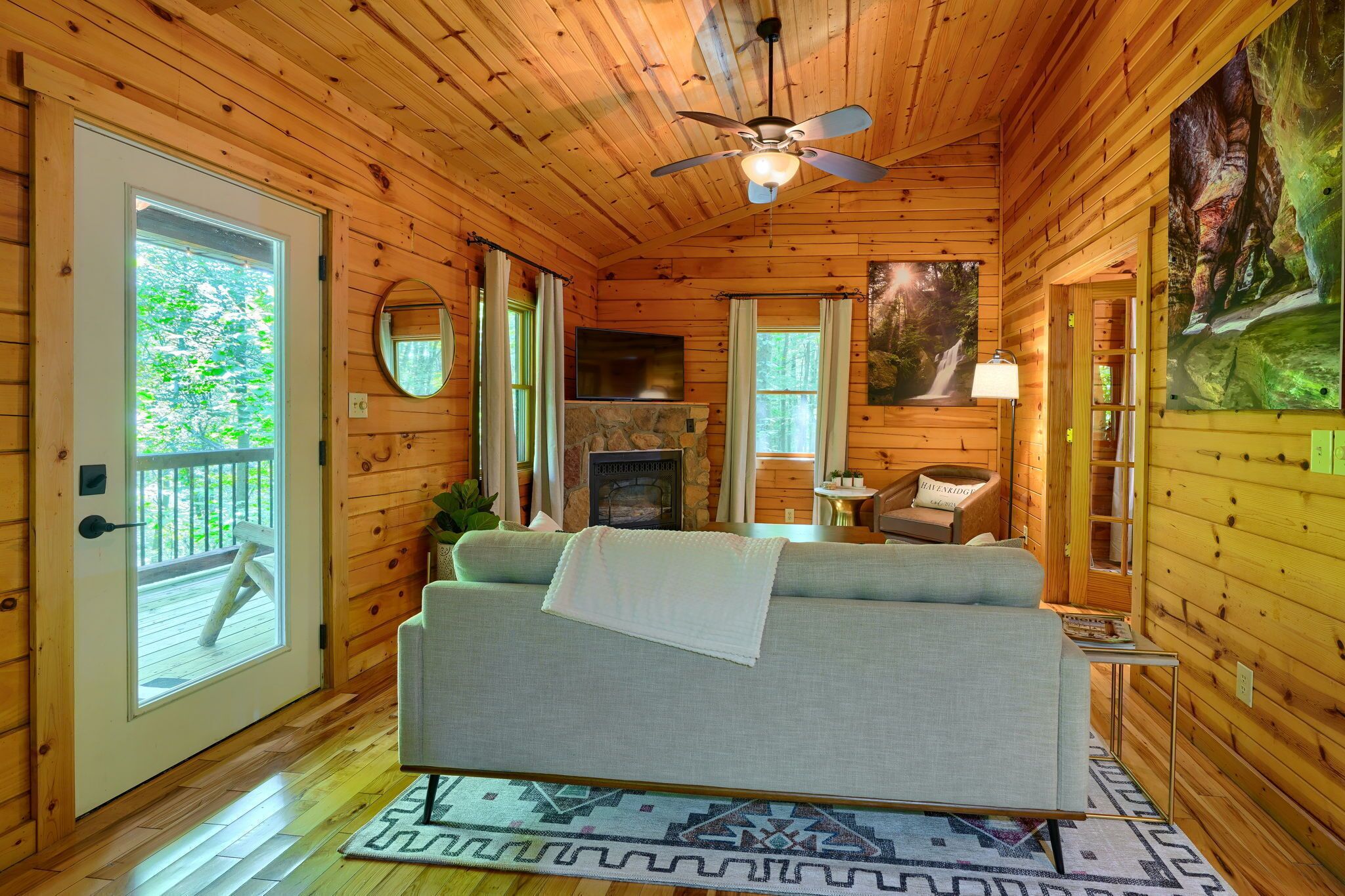 A cozy cabin interior with a fireplace, a sofa, a ceiling fan, and a large window.