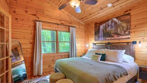 Une chambre dans une cabane en bois, avec un lit, un ventilateur de plafond, une fenêtre avec des rideaux et un tableau représentant une scène de forêt.