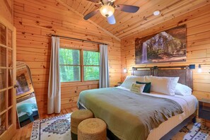A wooden cabin bedroom with a bed, a ceiling fan, a window with curtains, and a painting of a forest scene.