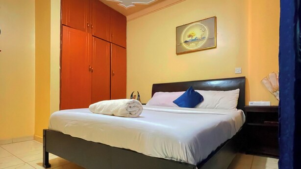 A neatly made bed with white linens and a blue pillow in a room with a tiled floor and a wooden wardrobe. - Rubee Homes (Kampala)