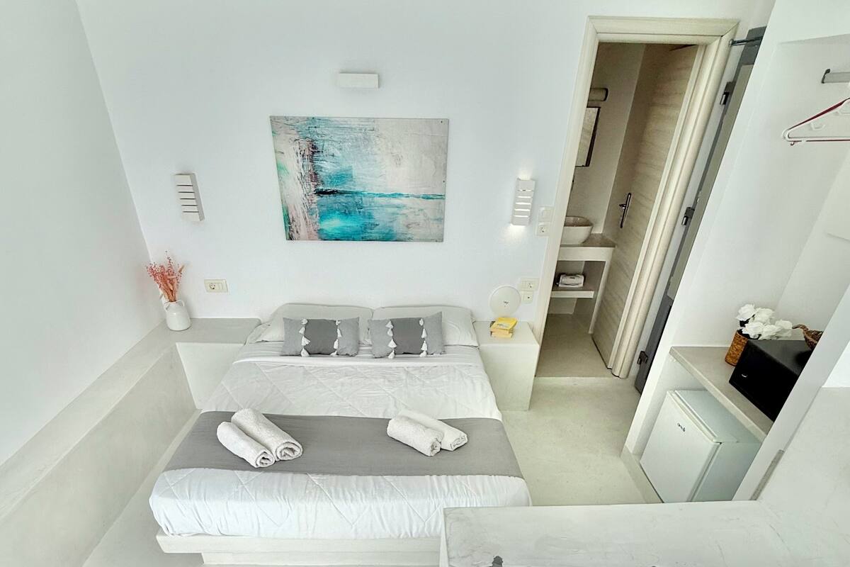 A modern hotel room with a large bed, a painting above it, a bathroom with a bathtub, and a view of the sea.