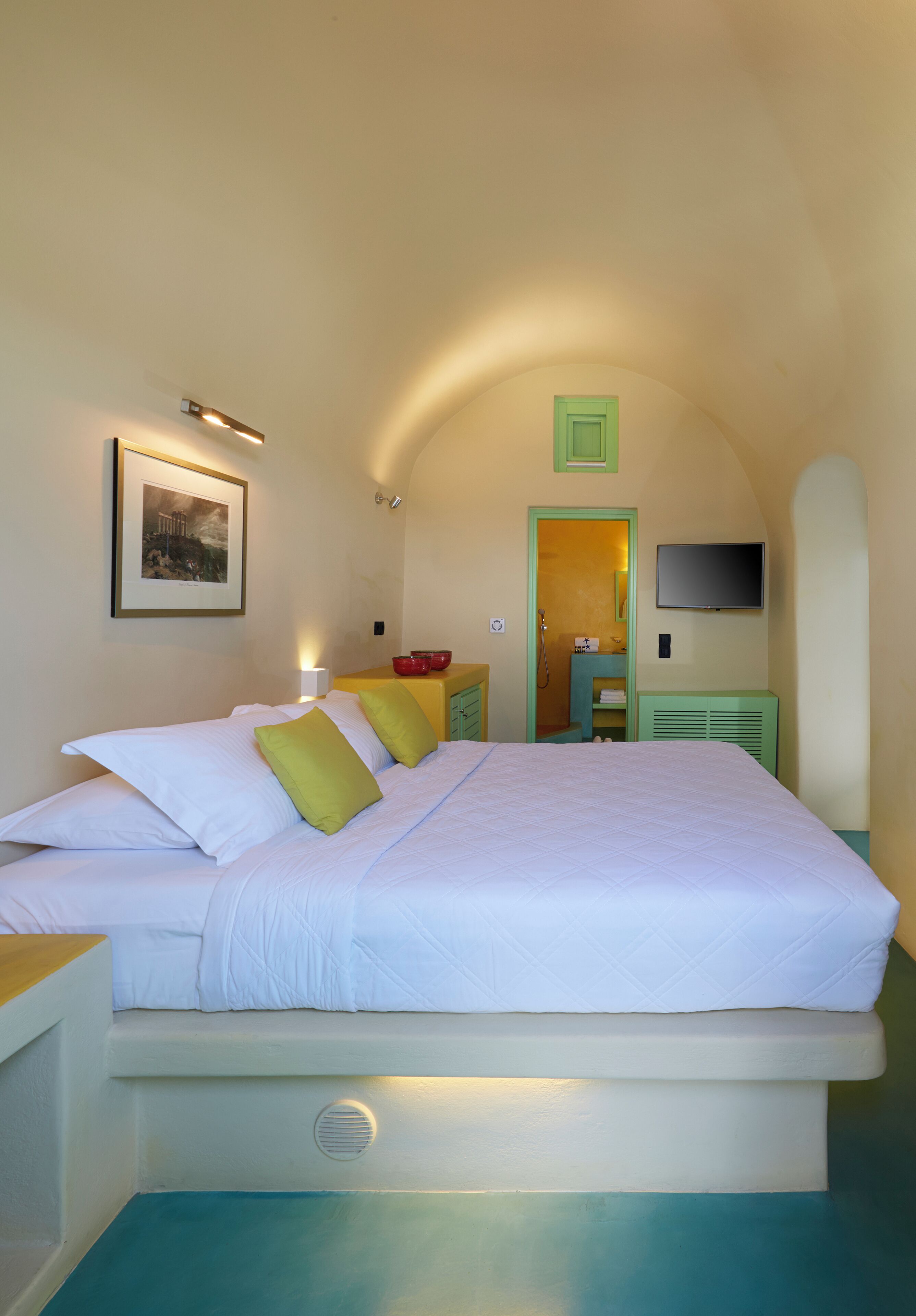 Un dormitorio con una cama, almohadas, una mesita de noche, un televisor montado en la pared y una puerta que da al baño.