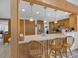 A modern kitchen with wooden cabinets, a central island with chairs, pendant lights, and a visible refrigerator.