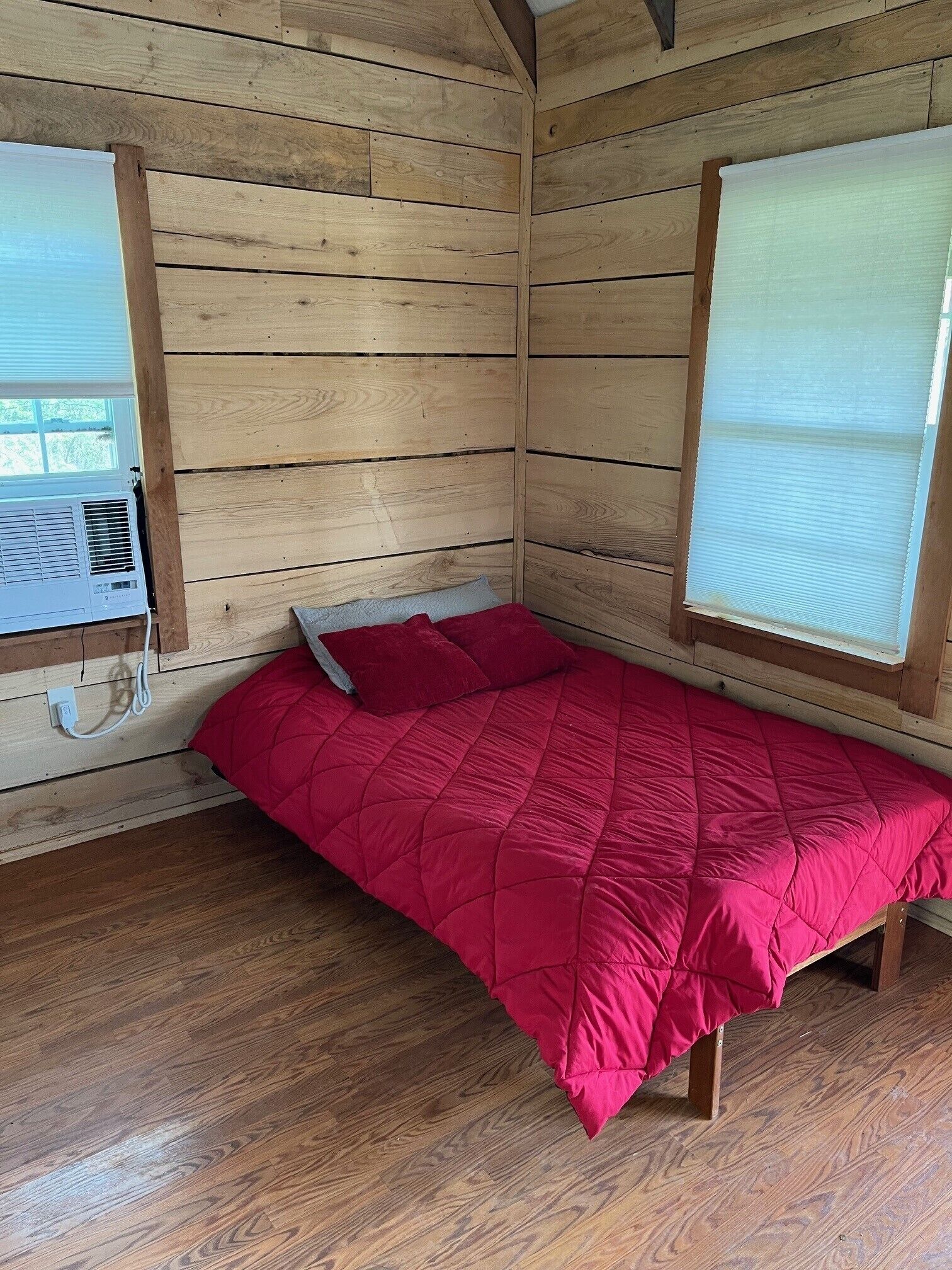 Une petite pièce chaleureuse aux murs en bois, avec un lit simple recouvert d’une courtepointe rouge.