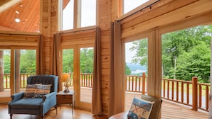 A wooden cabin interior with a blue armchair, a small table, and a leather bench with a cushion. Large windows offer views of a forest and a lake. - Eagle's Escape - Mountain top lodge with spectacular views (Huntingdon)