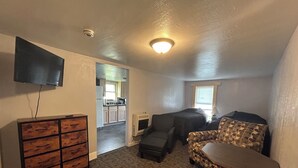 A compact living space with a kitchenette, a sofa, a patterned armchair, and a TV mounted on the wall.