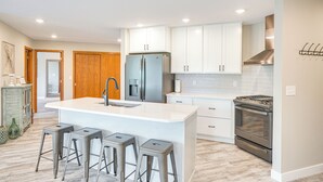 A modern kitchen with white cabinets, a stainless steel refrigerator, a built-in oven, and a central island with bar stools. - Family-Friendly ‘Lakeview Cottage’ w/ Deck & View (Yankton)