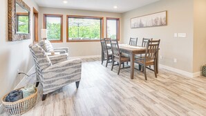 A dining area with a wooden table and chairs, large windows with views of greenery, a patterned armchair, and a woven basket on the floor. - Family-Friendly ‘Lakeview Cottage’ w/ Deck & View (Yankton)