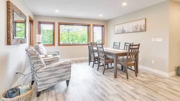 A dining area with a wooden table and chairs, large windows with views of greenery, a patterned armchair, and a woven basket on the floor.