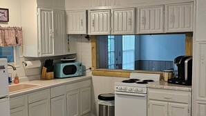 Una cocina compacta con armarios blancos, un microondas, estufa y un cubo de basura. Hay una mesa de madera con un libro y un control remoto.