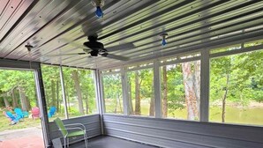 Un espacio exterior techado con suelo de hormigón, sillas de metal y vistas a árboles y una masa de agua.