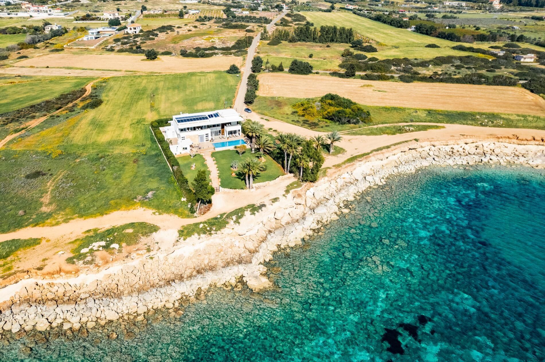 An aerial view of a coastal property with a large house, a swimming pool, and clear turquoise waters.