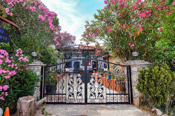 A gated entrance with a wrought iron gate, surrounded by lush greenery and pink flowers. - MADAM AFRODİT KONUKEVİ (Bozcaada)