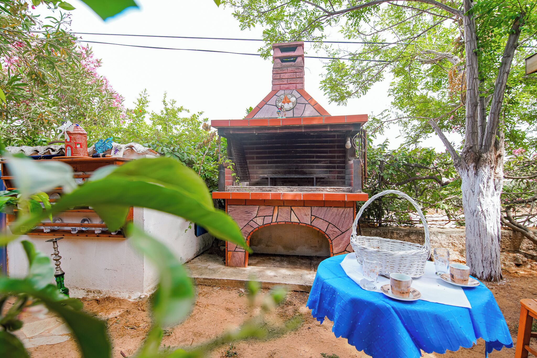 An outdoor stone oven with a chimney, surrounded by greenery and a table with cups and a basket.