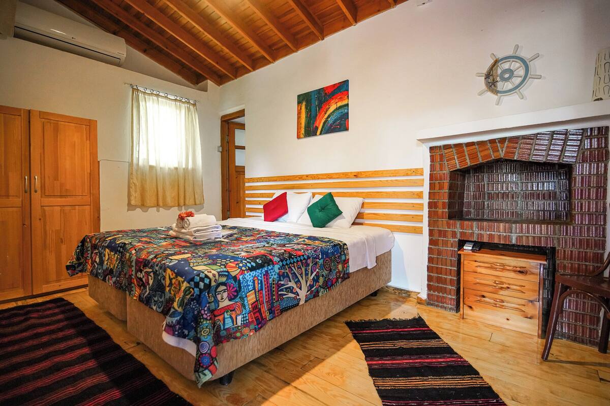 Quarto com cabeceira de madeira, colcha colorida, lareira e decoração de roda de navio na parede.