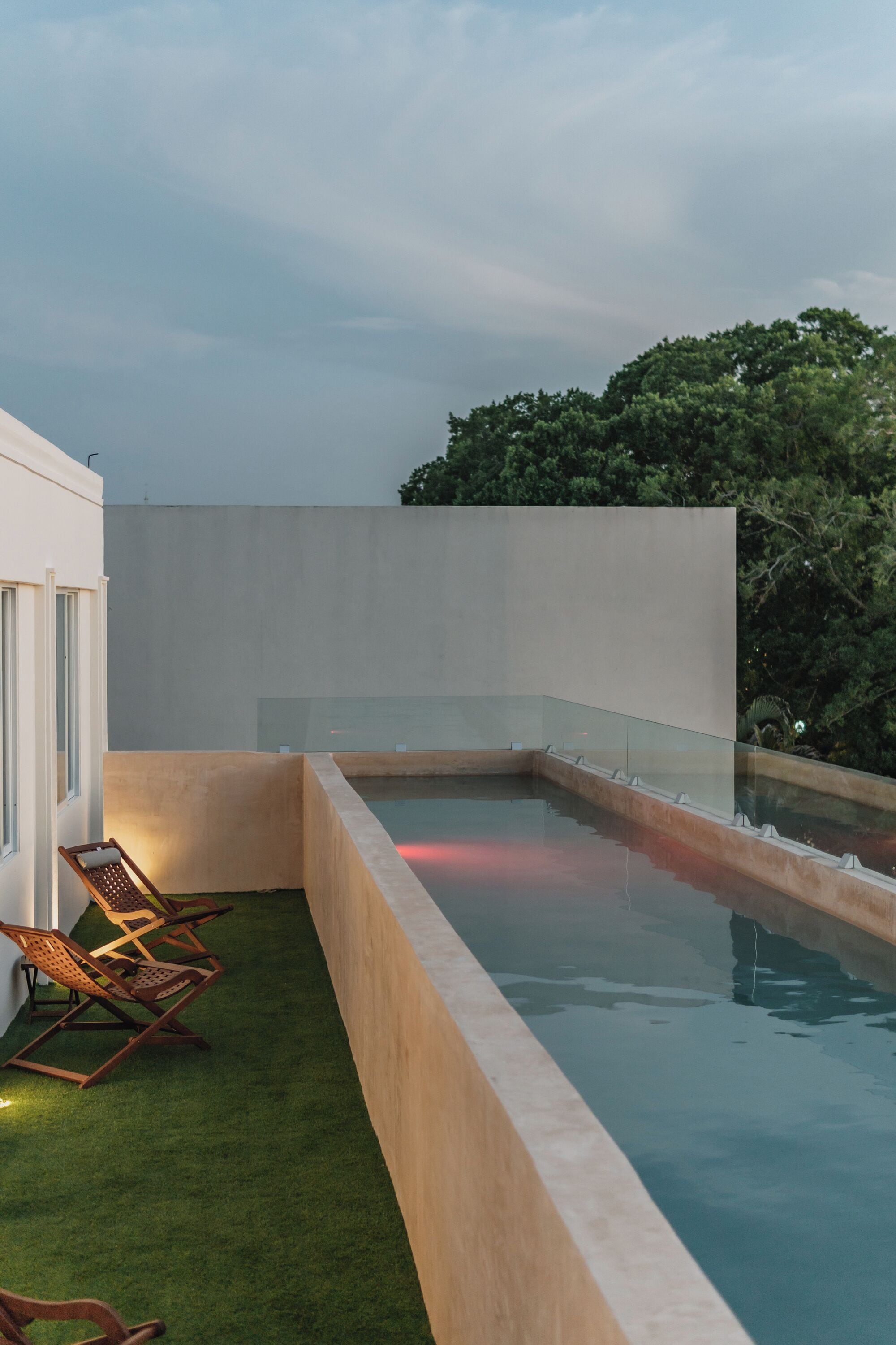 Una piscina en la azotea con barandilla de vidrio, rodeada por un muro bajo y un área verde con sillones de descanso.